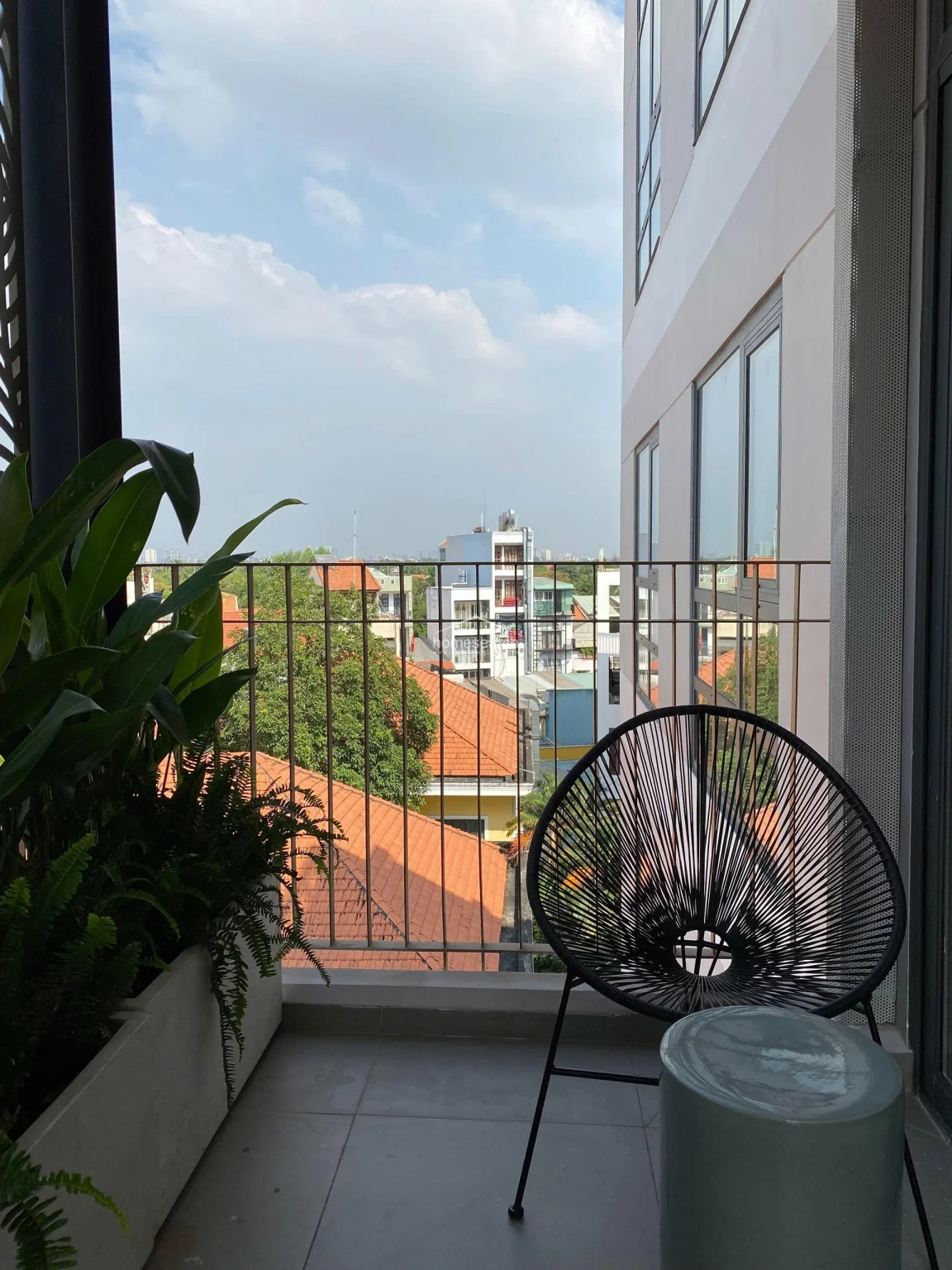 This cozy balcony provides a private outdoor retreat with a stylish black woven chair and a small side table, perfect for relaxing. The space is decorated with lush potted greenery and overlooks a quiet neighborhood with traditional red-tiled roofs and distant city buildings. The area is finished with neutral floor tiles and protected by a simple metal railing, offering an inviting transition between indoor and outdoor living.