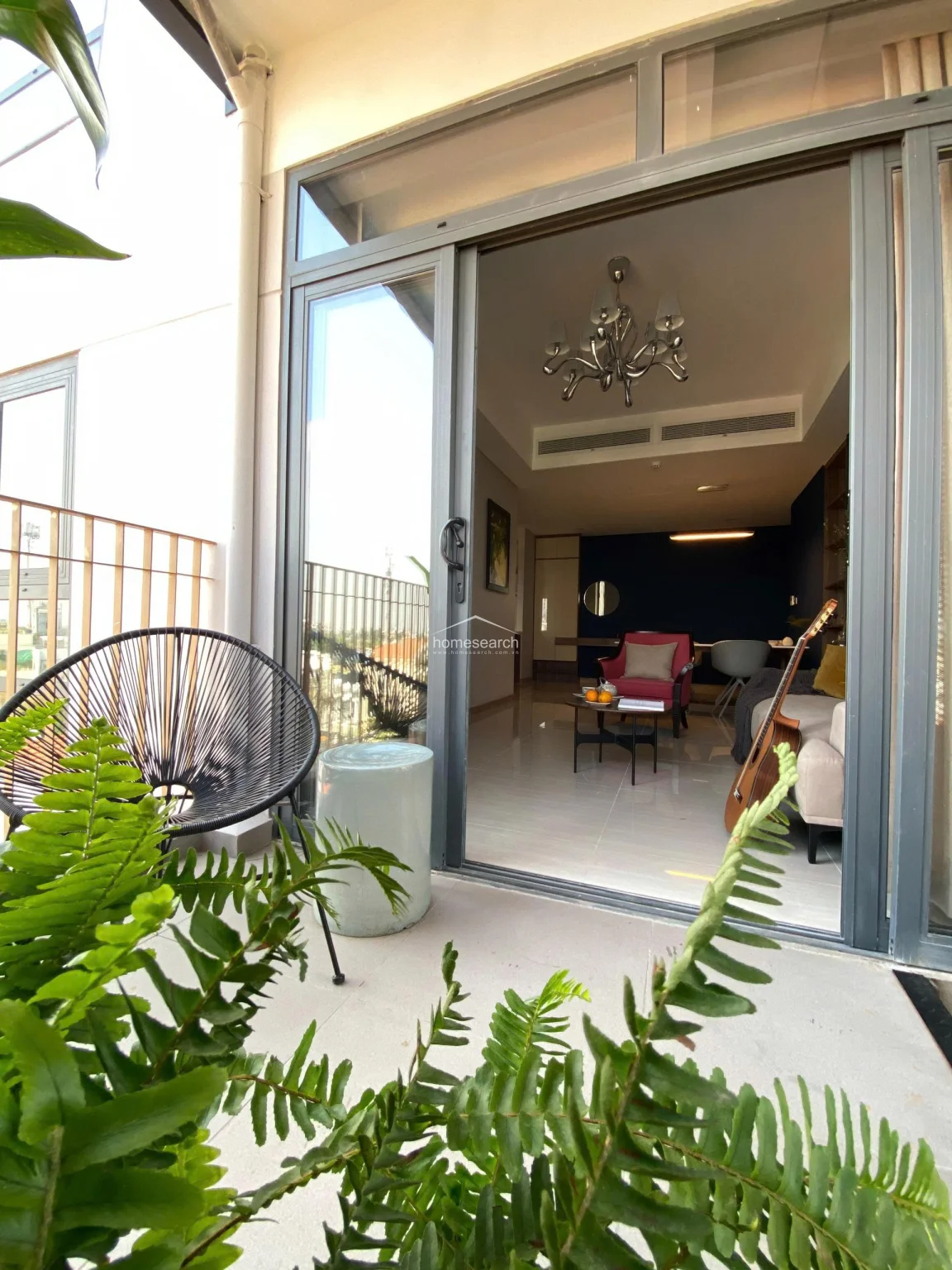 This view from the balcony highlights the indoor-outdoor connection, with a sliding glass door providing a clear sightline into the stylishly furnished living space. The balcony itself is a tranquil retreat, featuring a decorative metal privacy screen, a modern lounge chair, and lush potted greenery.
