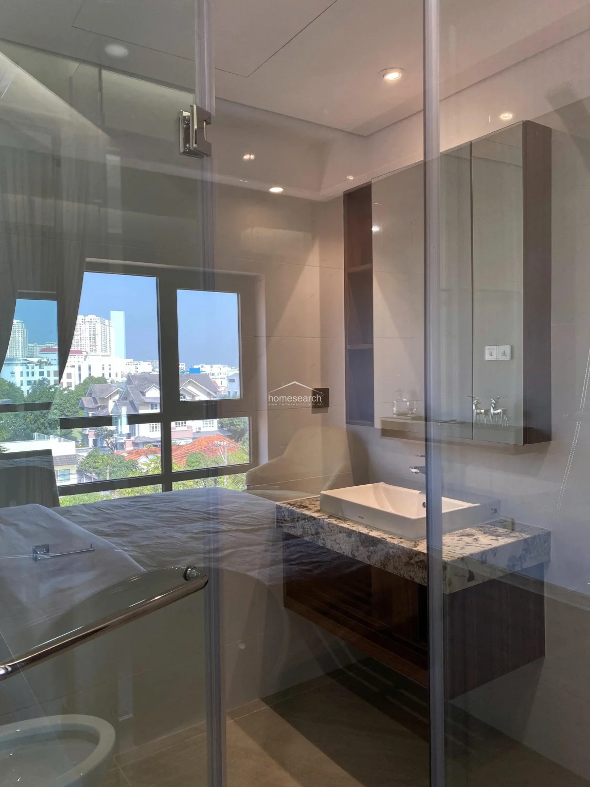 This modern bathroom features a sleek glass partition that offers a view directly into the adjacent bedroom, creating an open and airy atmosphere. The vanity includes a high-quality TOTO basin set on a speckled granite countertop with a dark wood slatted shelf below for storage. Recessed ceiling lighting and a large mirrored cabinet enhance the clean, contemporary aesthetic of the space.