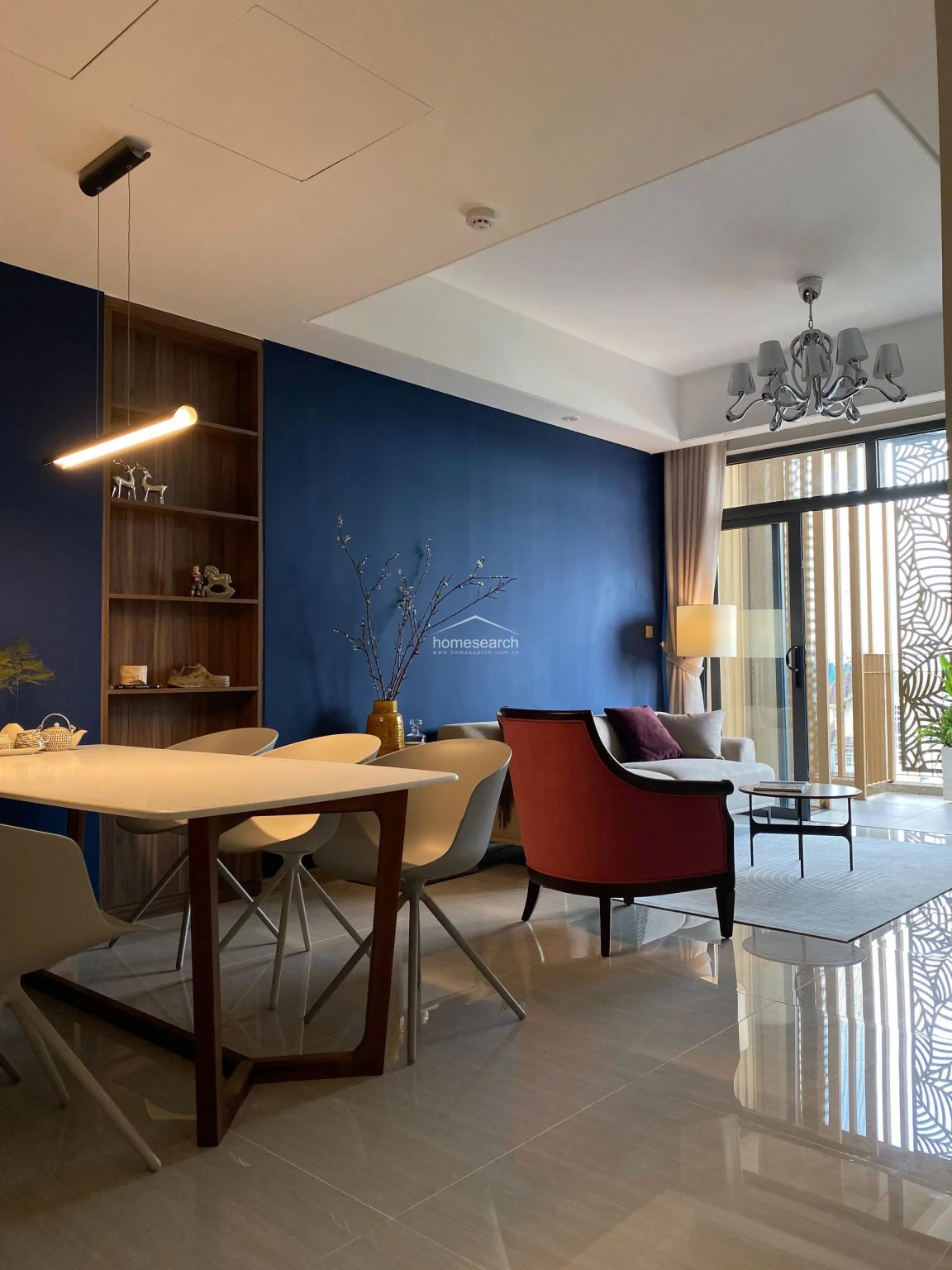The dining space is anchored by a contemporary white table and modern chairs, illuminated by a sleek, minimalist linear pendant light. The room features a tall, built-in wooden shelving unit set against a bold navy wall, creating a sophisticated transition between the dining zone and the living area.