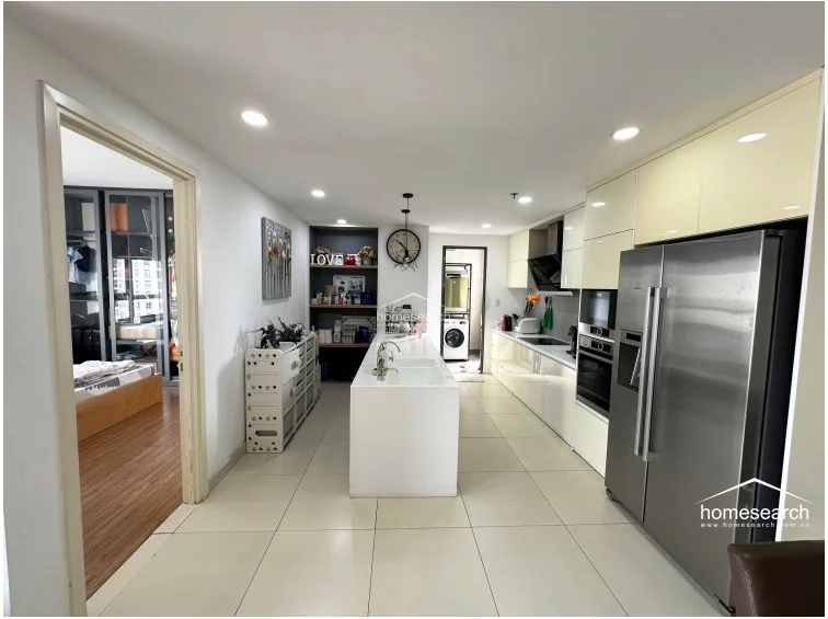 The kitchen is highly functional with a sleek, white-cabinet design, integrated stainless steel appliances including a double-door refrigerator, and a central island that serves as a breakfast bar. The space is bright and organized, connecting directly to the dining area and featuring recessed ceiling lighting for a clean, modern aesthetic.