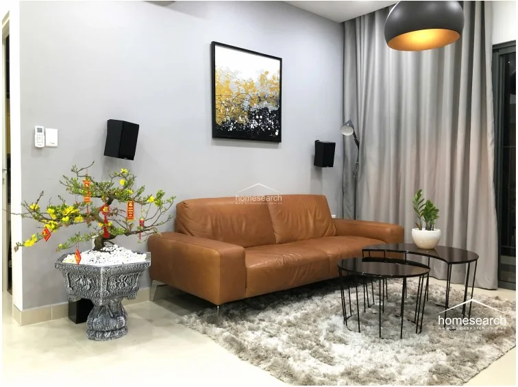 The living area is anchored by a comfortable tan leather sofa and a textured rug, creating a cozy focal point beneath a modern pendant light. The space is well-maintained and features a minimalist design with grey walls, a wall-mounted television, and integrated audio equipment.