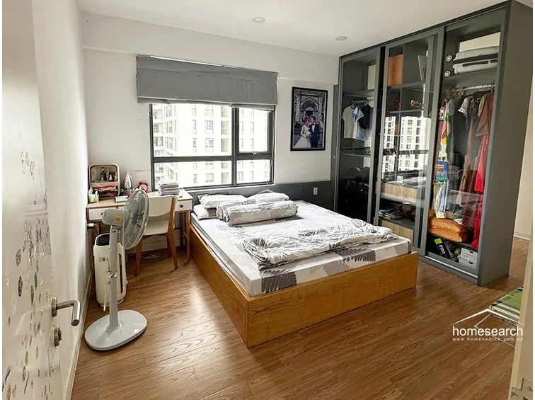 This bedroom features a warm wood-toned floor and a large, modern floor-to-ceiling wardrobe with glass-paneled doors, offering excellent visibility and storage. A large window with a grey Roman shade provides natural light and an urban view, while the space is efficiently furnished with a bed, a compact desk, and a portable fan. The room is in good condition, characterized by a neutral color palette and clean, contemporary lines.