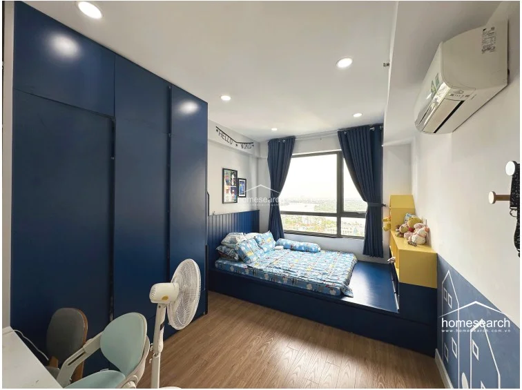 This child-friendly bedroom is designed with a vibrant blue and white color scheme, featuring a built-in platform bed and an extensive floor-to-ceiling navy storage unit. A large window draped in blue curtains offers a scenic elevated view, and the room is equipped with a wall-mounted AC unit and recessed ceiling lights. The space is well-maintained, utilizing wood-look flooring and custom cabinetry to maximize functionality in a compact footprint.