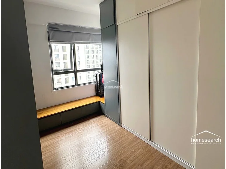 This versatile room serves as a functional storage or multi-purpose space, featuring a sleek, floor-to-ceiling sliding-door wardrobe in a mix of white and dark grey finishes. A custom-built wooden bench with integrated lower cabinets runs along the window, providing both seating and additional storage options. The room maintains a consistent aesthetic with wood-look flooring and a clean, minimalist design, appearing to be in excellent condition.