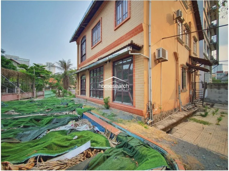 This exterior view shows a multi-story villa with a classic brick-and-stucco facade and a prominent pitched roof. The property features large, arched windows and an integrated balcony, though the surrounding grounds are currently unkempt and covered with protective green mesh.