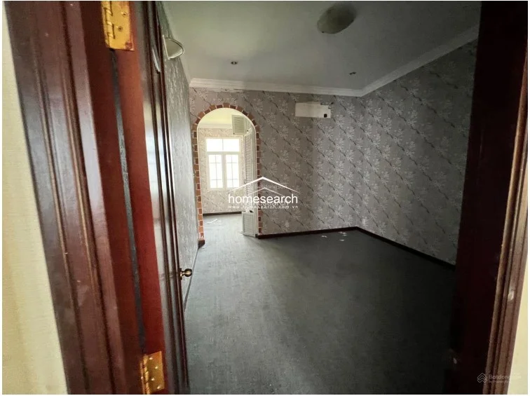 This bedroom or secondary living space is finished with grey patterned wallpaper and grey carpeting, creating a distinct aesthetic from the other rooms. An arched doorway leads to an adjoining space, and the room includes a wall-mounted AC unit, indicating a functional layout that requires some cosmetic refreshing.