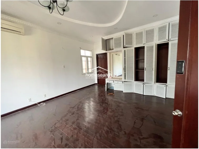 This spacious bedroom features dark hardwood flooring and white walls, complemented by a large, built-in white louvered wardrobe system with integrated vanity storage. The room is equipped with a wall-mounted AC unit and receives natural light from a window, while a decorative ceiling cove adds an architectural touch.