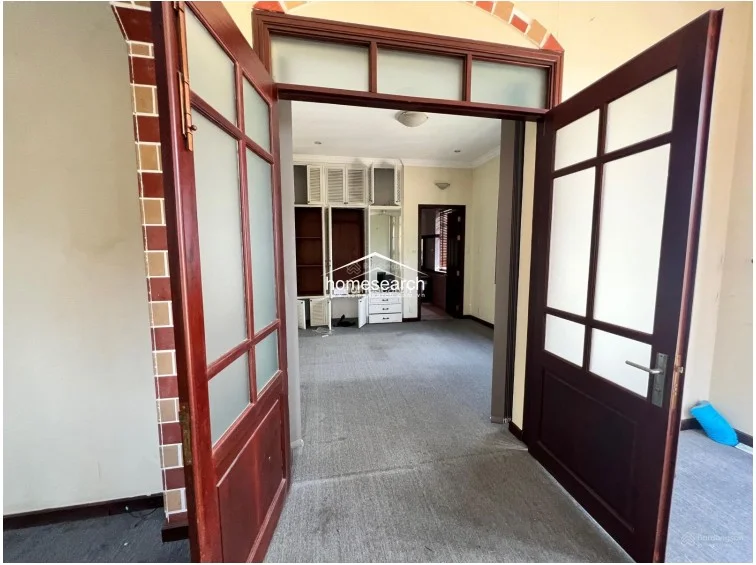 This entryway or transition space is framed by a decorative brick-accented archway and features a set of elegant, multi-pane glass and wood doors. The interior beyond shows a carpeted room with a large, white built-in wardrobe and vanity unit, suggesting a functional dressing or bedroom layout.