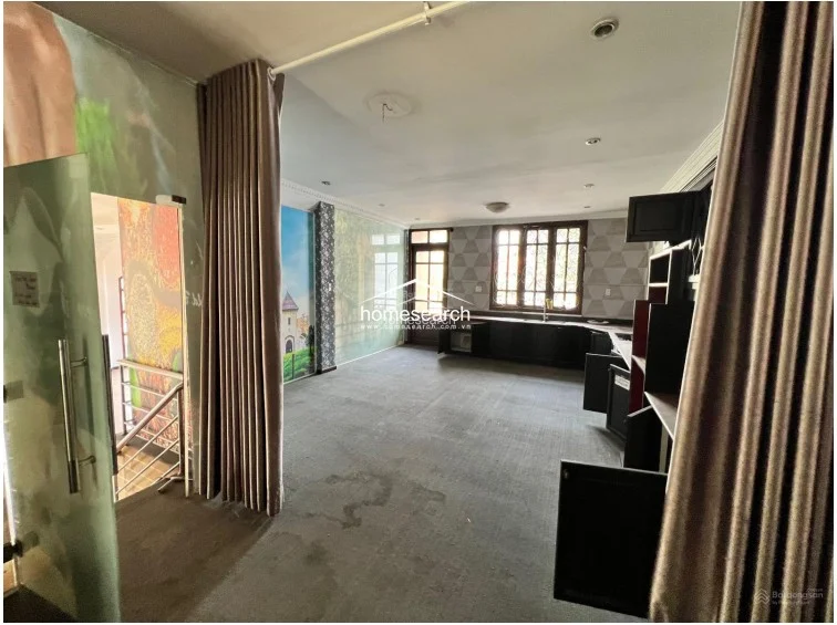 This expansive room serves as a multi-functional space with grey carpeting and unique, mural-style wall coverings that add a distinct decorative element. It includes a dark wood kitchen cabinetry section, a large window with security bars, and heavy floor-to-ceiling curtains that provide privacy and partition the area from the stairwell.