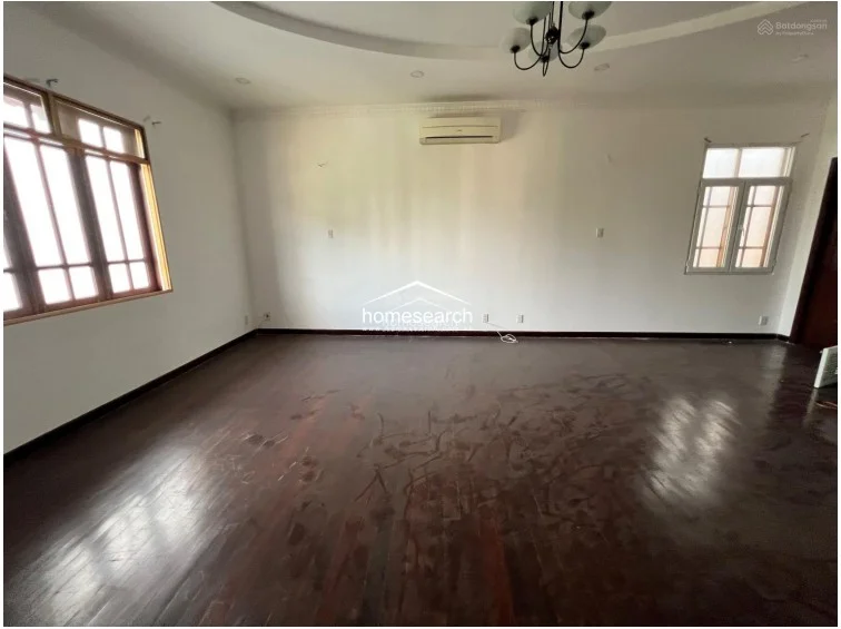 This spacious living or primary room showcases rich dark wood flooring and a decorative recessed ceiling with a classic chandelier, creating an elegant atmosphere. Large windows provide ample natural light, while the neutral white walls and wall-mounted air conditioning unit offer a clean, functional backdrop for future furnishings.