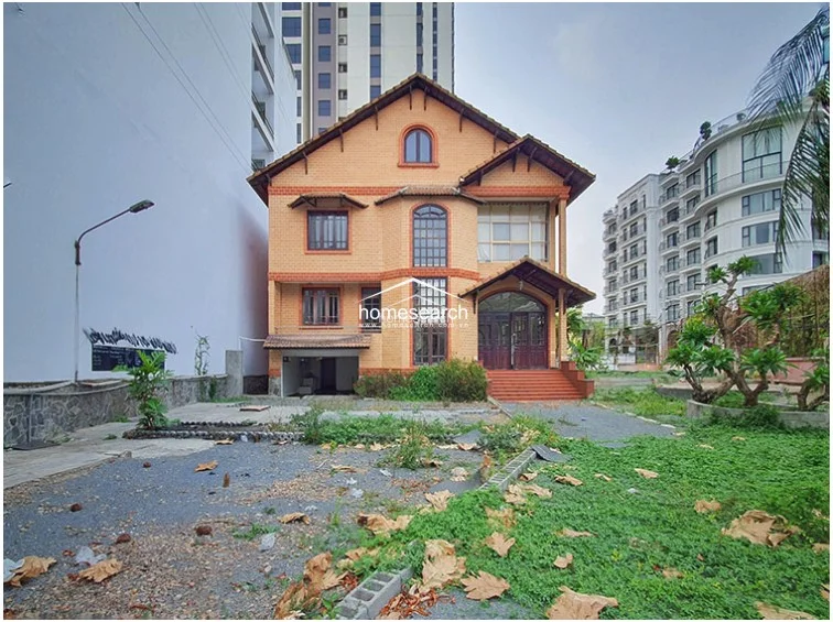 This angle highlights the side of the villa, emphasizing the exterior brickwork and the placement of several wall-mounted AC units. The ground area is partially paved with tiles, but the overall maintenance level appears to be in need of attention due to debris and overgrown vegetation.
