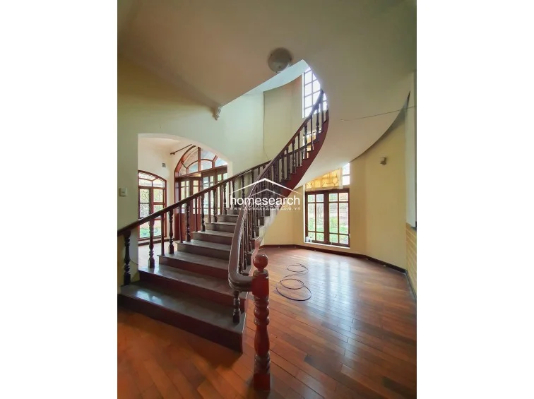 This interior perspective shows a grand foyer featuring a sweeping wooden spiral staircase that serves as the home's focal point. The space is finished with warm-toned hardwood flooring and pale yellow walls, creating an open, airy flow that connects to the adjacent living areas.