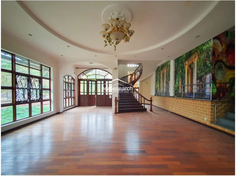 This expansive living area features a sophisticated open-plan layout with a coffered ceiling, a decorative chandelier, and a unique wall mural. The room is flooded with natural light from the large, grid-patterned windows and connects directly to the central staircase, showcasing a blend of traditional architectural details and spacious design.