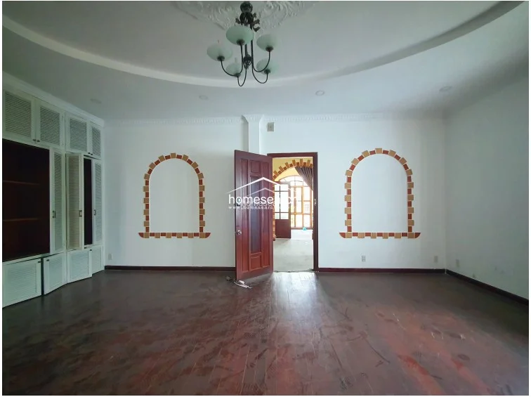 This bedroom features warm hardwood flooring, white walls, and a recessed ceiling design with a decorative chandelier. It includes extensive built-in white louvered cabinetry, a matching vanity, and a wall-mounted air conditioning unit, presenting a clean but slightly dated aesthetic.