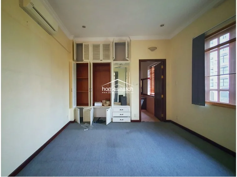 This bedroom features a practical layout with wall-to-wall carpeting and a large, built-in white louvered wardrobe unit that includes a central vanity area. The room is illuminated by natural light from a side window and is equipped with a wall-mounted air conditioning unit, though the overall condition appears to need some cosmetic refreshing.