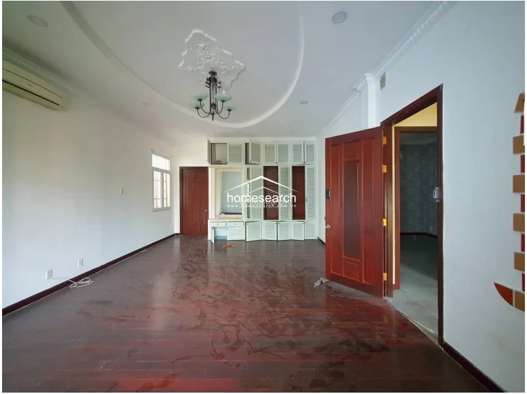 This bedroom space maintains consistency with the rest of the home, featuring hardwood floors, white walls, and a central ceiling medallion with a chandelier. The room is equipped with a wall-mounted AC unit and provides a clear view into an adjacent room through a wooden door, highlighting a functional, open-flow layout.