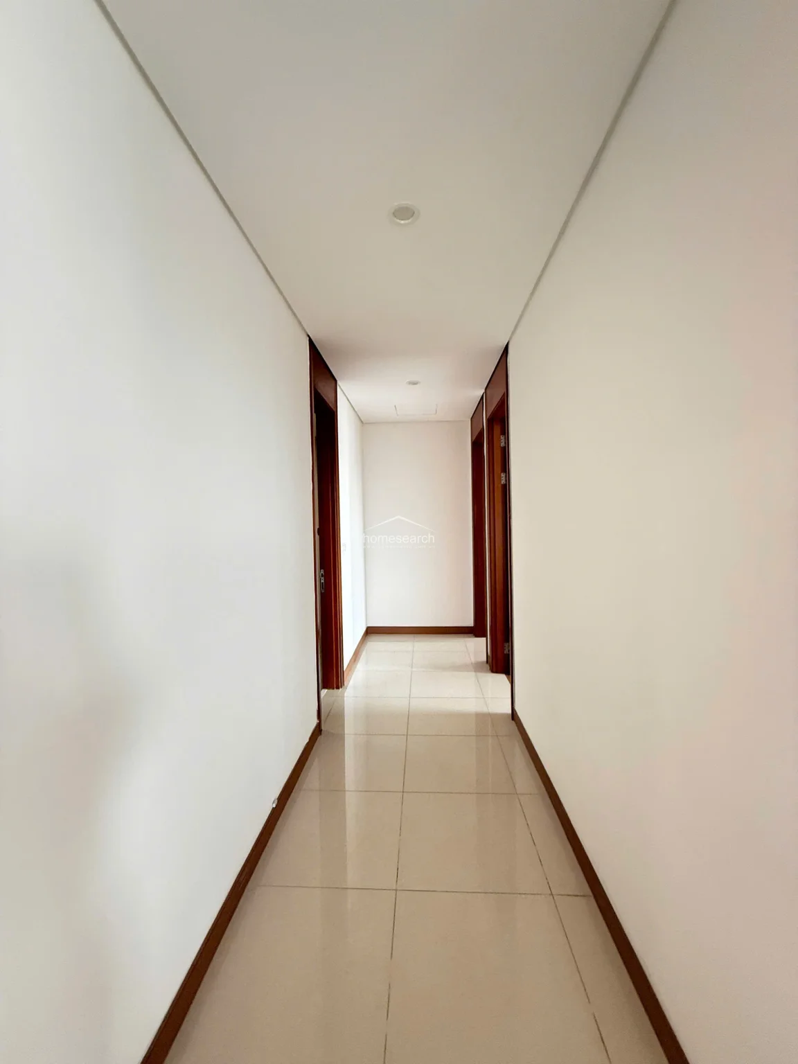 This interior hallway features clean white walls and light-colored square floor tiles, creating a bright and airy transition space. The corridor is framed by dark wood door casings that provide a sharp contrast, leading to the various rooms of the property.