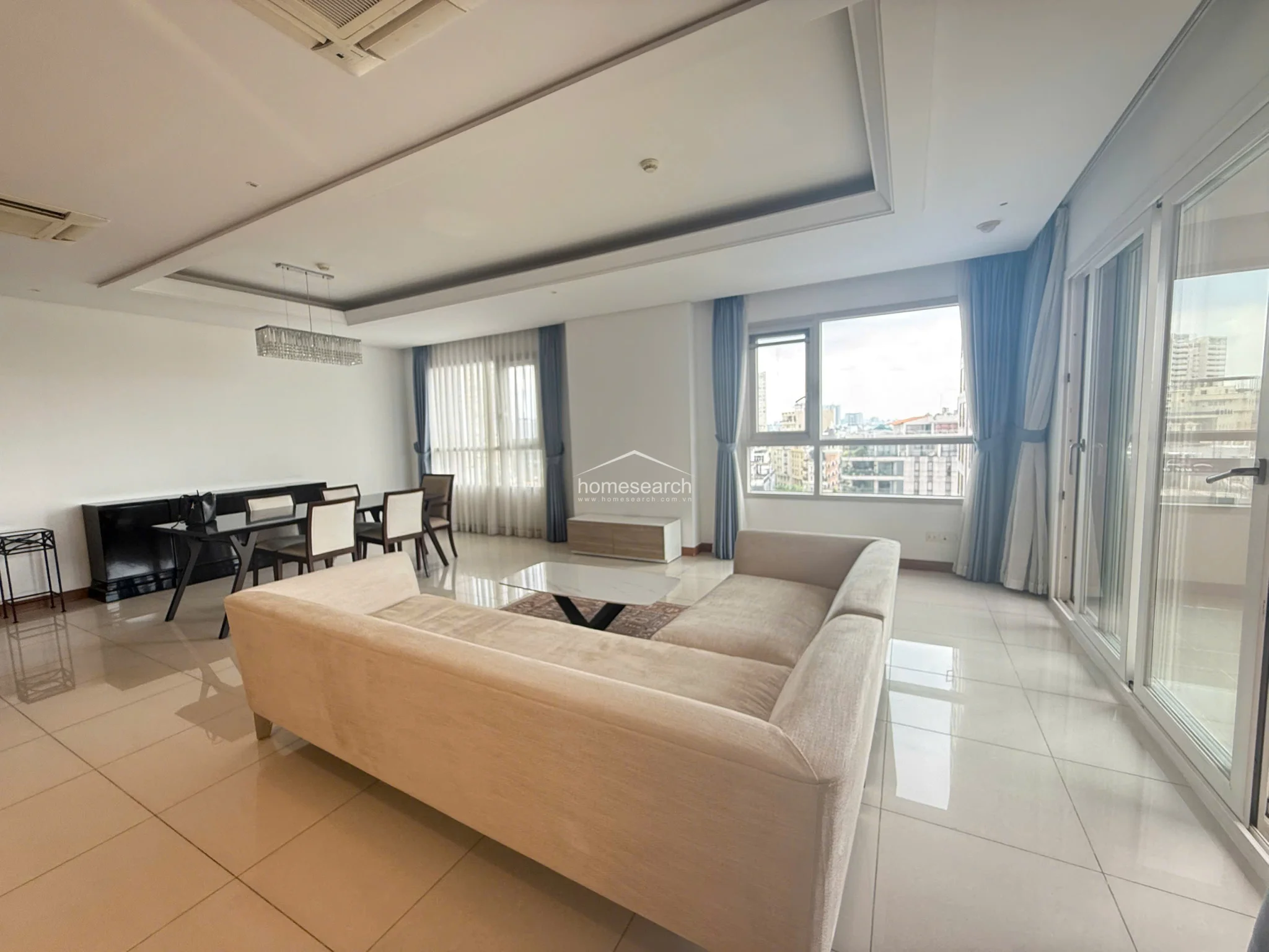 This open-concept living and dining area features a bright, airy layout with large windows that provide ample natural light and an expansive city view. The space is finished with polished light-colored floor tiles, white walls, and a modern tray ceiling with recessed lighting and a crystal chandelier. The room is well-maintained and includes a neutral-toned sectional sofa, a glass-top coffee table, and a contemporary dining set.