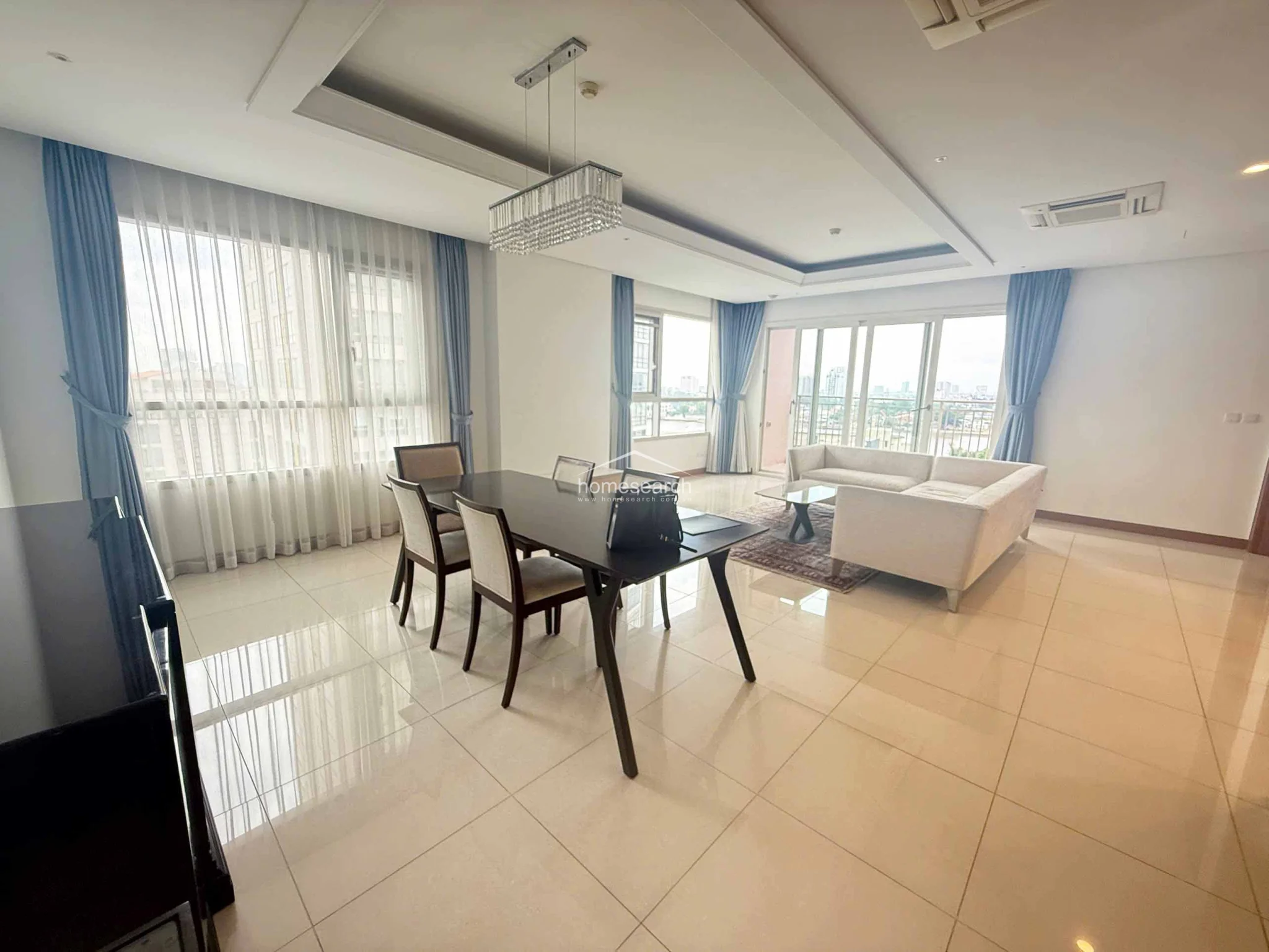 The dining area is seamlessly integrated into the main living space, creating a fluid transition between zones. It is anchored by a sophisticated crystal chandelier and large windows draped in elegant floor-to-ceiling curtains. The neutral color palette and polished tile flooring contribute to a clean, minimalist aesthetic throughout the room.