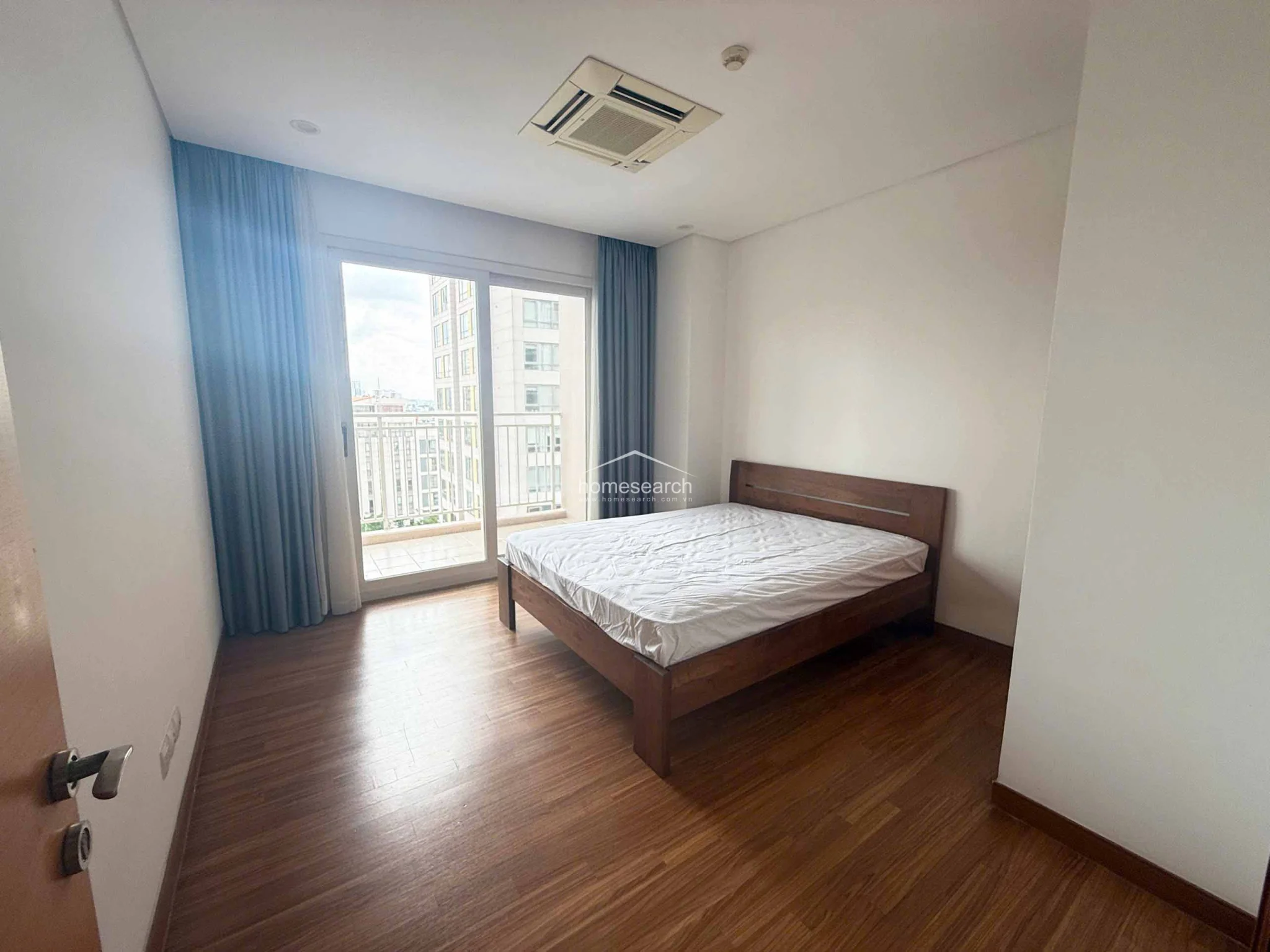 This bedroom offers a minimalist aesthetic with warm wood flooring, white walls, and a simple wooden bed frame. A sliding glass door provides direct access to a private balcony, while a ceiling-mounted AC unit ensures comfort in the space.