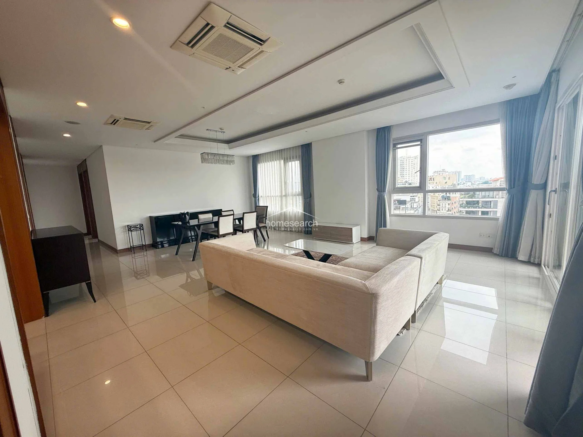 The living room layout is spacious and functional, centered around a large sectional sofa and a patterned area rug that adds texture to the space. Large windows and glass doors allow for significant natural light, while the tray ceiling and recessed lighting add architectural interest. The room is in pristine condition, offering a comfortable and stylish environment.