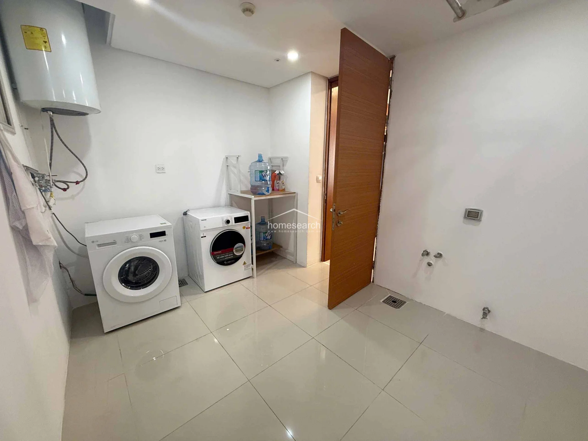 This functional laundry and utility room features clean white walls and polished light-colored tile flooring, providing a bright and hygienic workspace. It is equipped with two front-loading washing machines, a wall-mounted water heater, and an open shelving unit for storage, with additional plumbing hookups available on the adjacent wall. The space connects directly to the rest of the home via a wooden door and appears well-maintained with a minimalist, utilitarian layout.