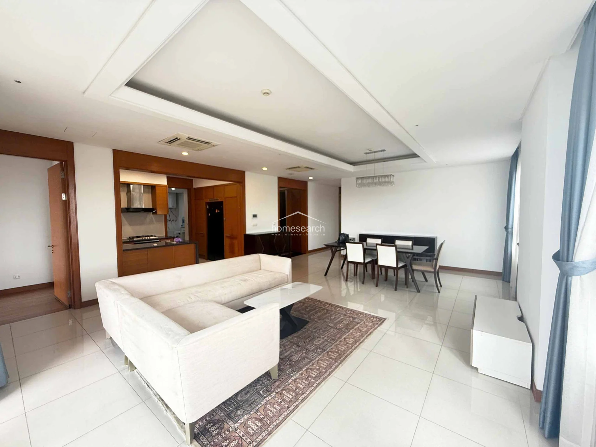 This view highlights the connection between the living area and the adjacent kitchen, which is framed by warm wood-paneled accents. The room maintains a consistent, high-end feel with its neutral furniture, polished tile flooring, and integrated ceiling-mounted AC units. The overall condition is excellent, reflecting a modern, well-kept interior design.