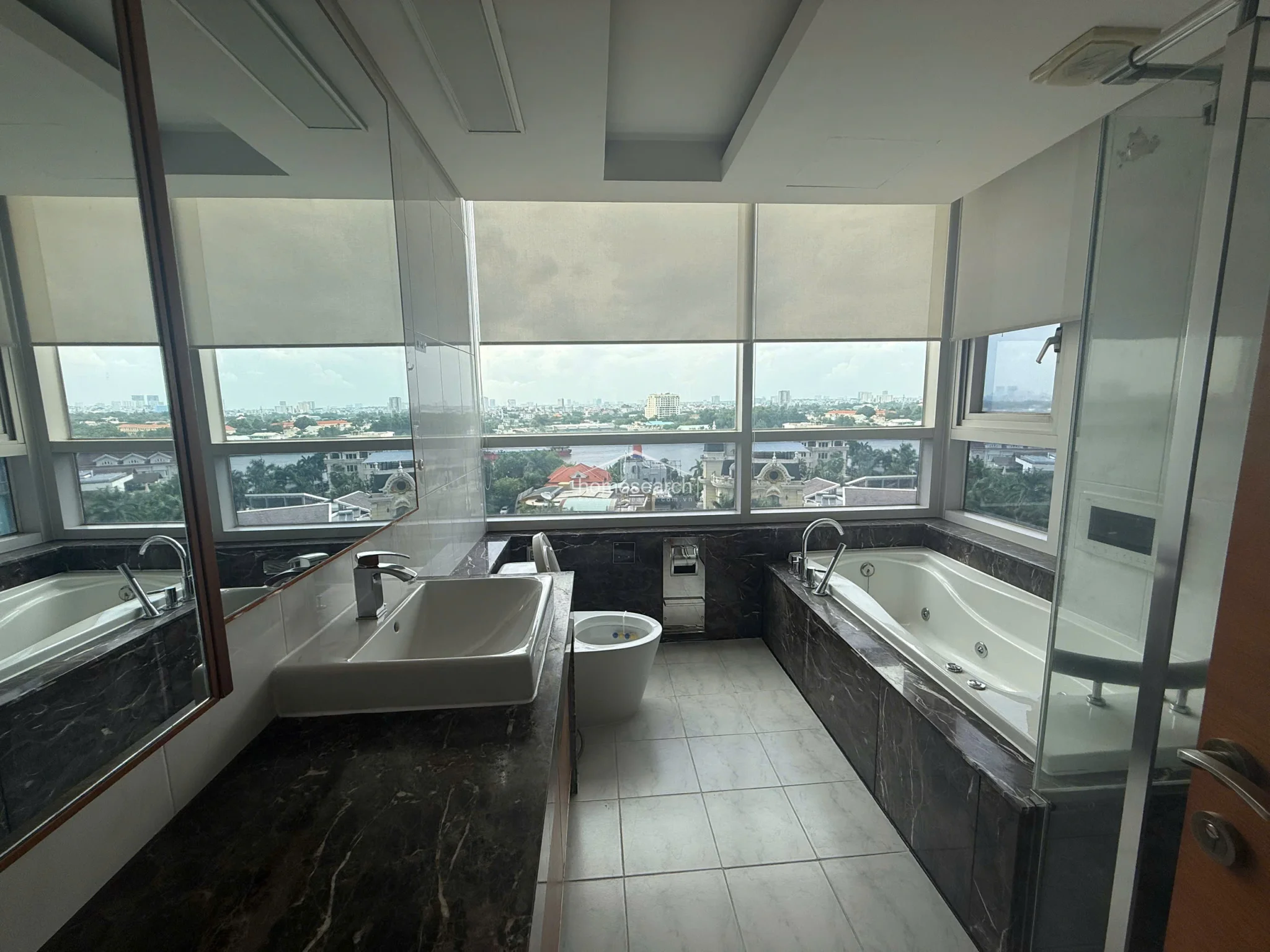 This modern bathroom is designed with a luxurious dark marble vanity and matching tub surround, complemented by light-colored floor tiles. It features a large window with roller shades that provides a panoramic city view, a separate glass-enclosed shower area, and a sleek, contemporary sink.