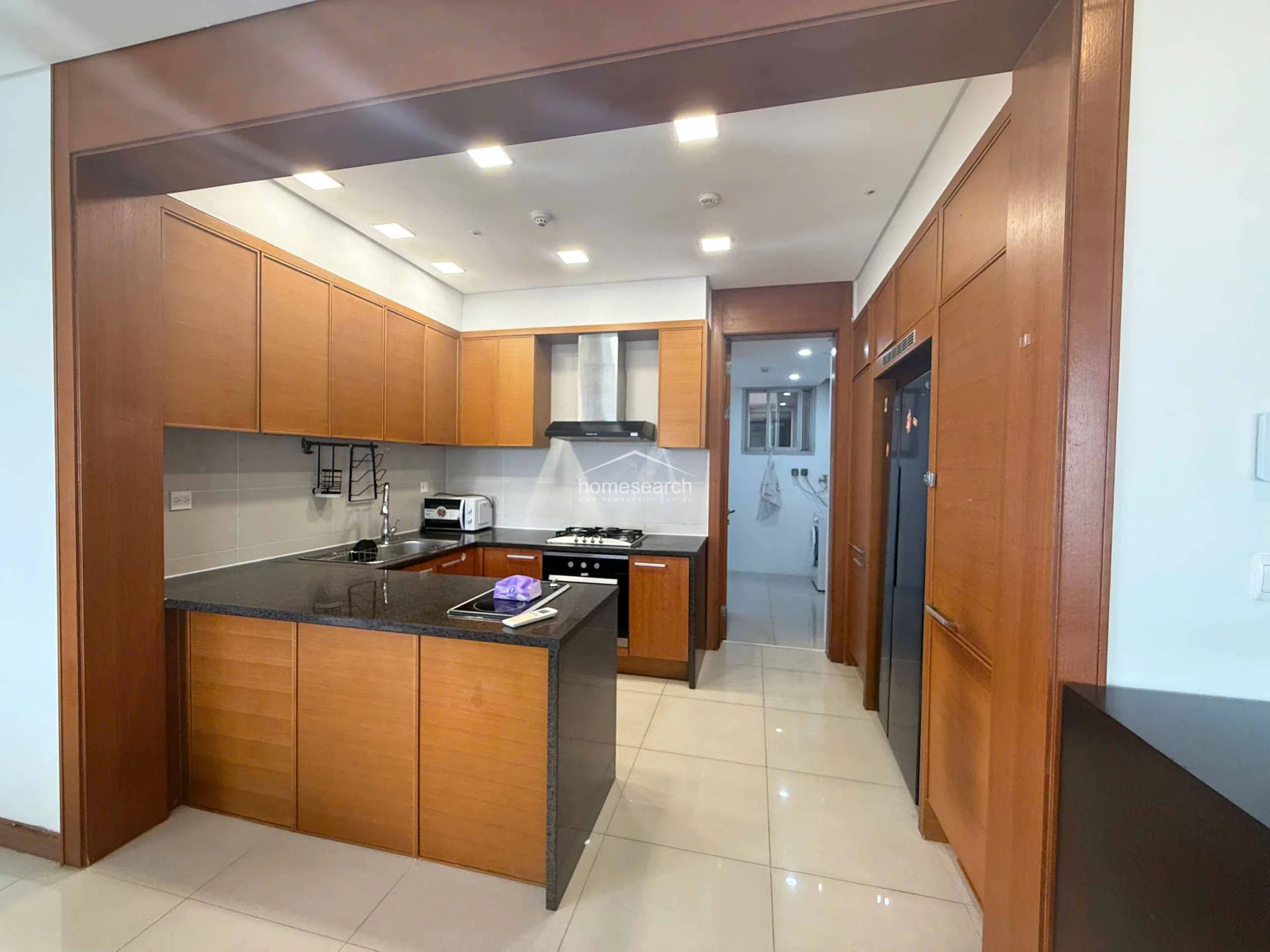 The kitchen features a functional U-shaped layout with warm-toned wooden cabinetry and contrasting dark granite countertops. It is fully equipped with a built-in oven, gas stove, stainless steel range hood, and a large black refrigerator. A doorway leads to a utility area, and the space is finished with the same polished tile flooring found throughout the home, ensuring a cohesive design.