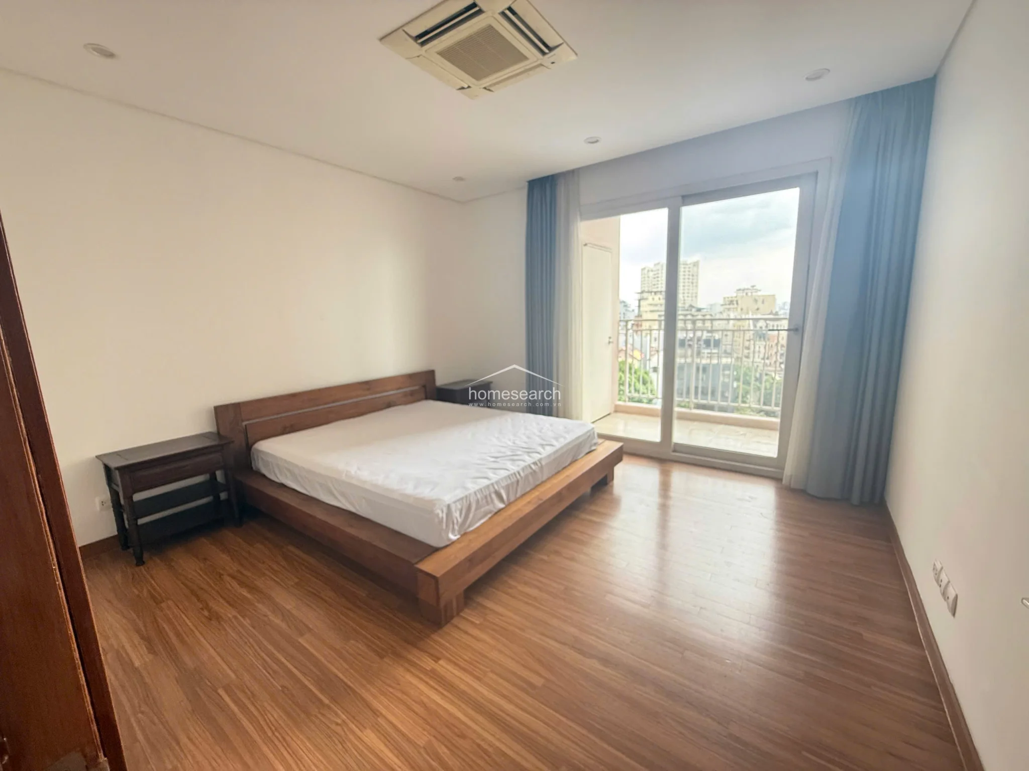 This bedroom maintains a consistent design language with wood flooring and a clean, neutral color palette. It is furnished with a wooden bed frame and side tables, and includes a sliding door leading to an outdoor balcony with city views.
