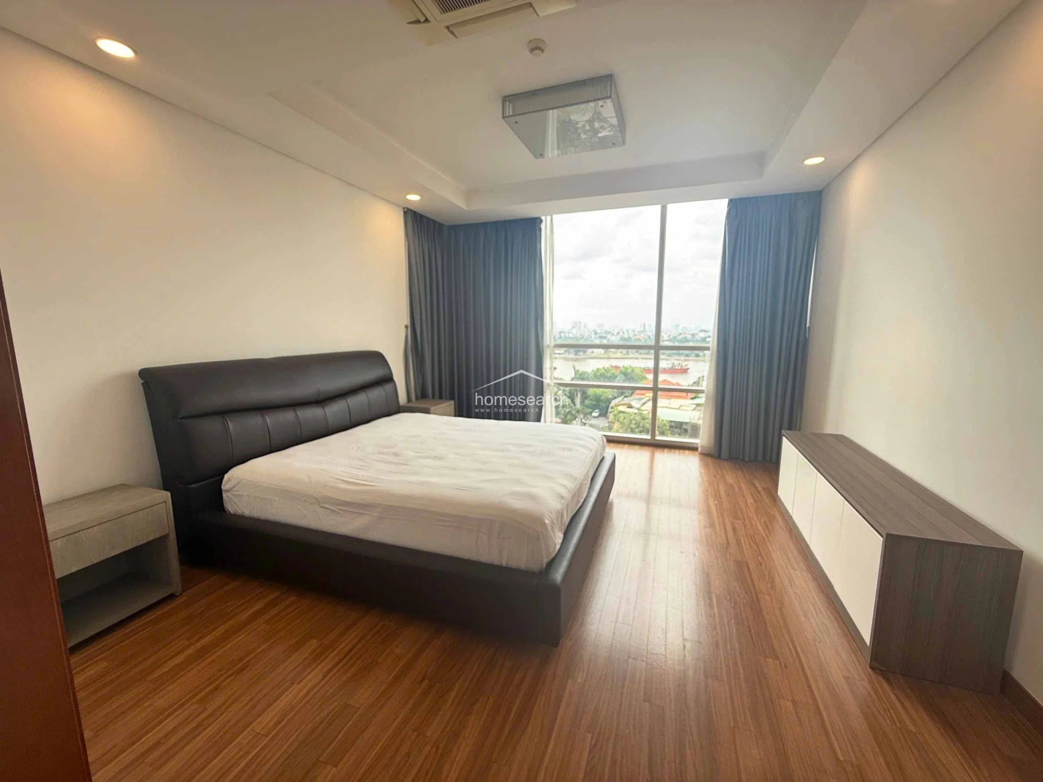 This primary bedroom features warm wood flooring and a large floor-to-ceiling window that offers an expansive city and river view. The room is furnished with a dark leather-upholstered bed, a matching low-profile media console, and includes a ceiling-mounted AC unit for climate control.