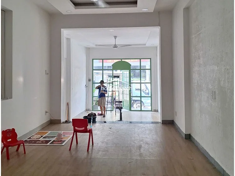 This perspective looks toward the front entrance, highlighting a large, green-framed glass door system that allows for significant natural light. The interior maintains a minimalist aesthetic with white walls, wood-look flooring, and a ceiling fan, providing a clean and open flow throughout the ground floor.