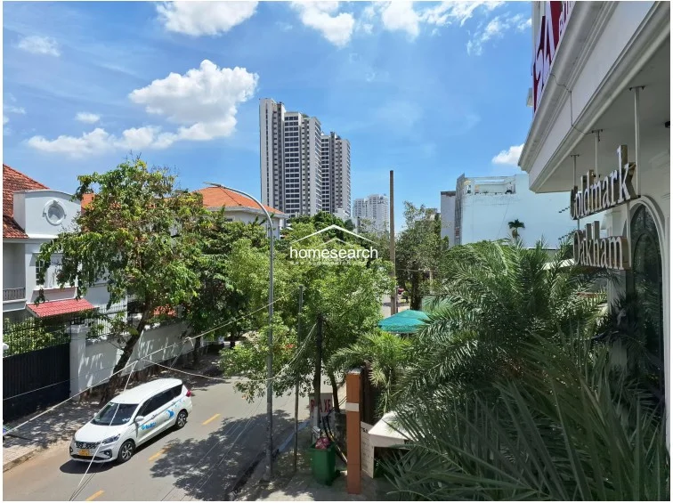 This outdoor view from the building balcony showcases an urban streetscape with lush greenery and a clear line of sight to high-rise residential towers. The perspective highlights the building's exterior architecture, featuring white facades and arched window detailing, suggesting a well-maintained property in a developed neighborhood.