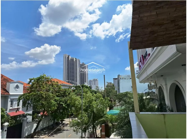This perspective is similar to the previous shot, emphasizing the building's architectural character with its white exterior, arched design elements, and wooden-look soffit detailing on the balcony. The view captures a pleasant mix of mature trees and modern city infrastructure under a bright, partly cloudy sky.