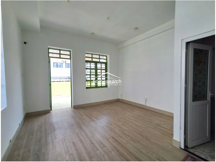 This interior space is a clean, minimalist room featuring light wood-grain laminate flooring and crisp white walls, providing a bright and neutral canvas. The room includes a direct access point to an outdoor balcony through a green-framed door, and a matching window that allows for good natural light and airflow.