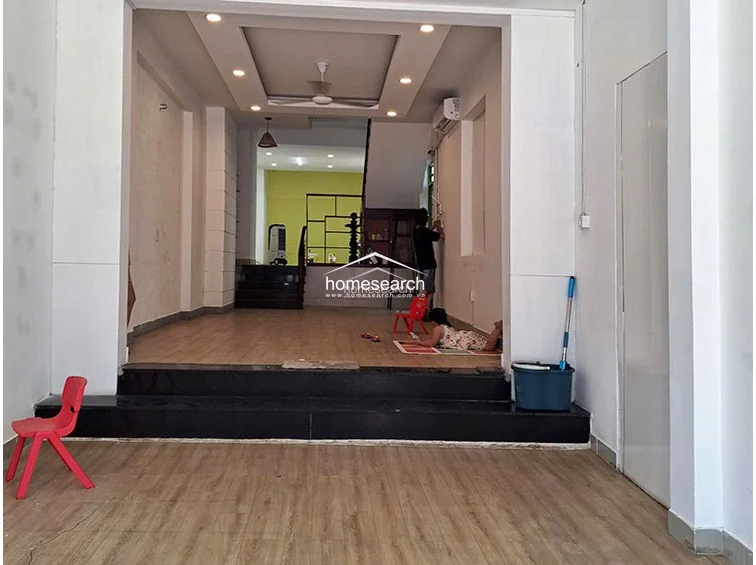This image shows a multi-level open-plan living area featuring light-toned wood-look flooring and a distinct transition created by a black stone step. The space is illuminated by recessed ceiling lights and a central fan, with white walls and a decorative textured pillar adding architectural interest.
