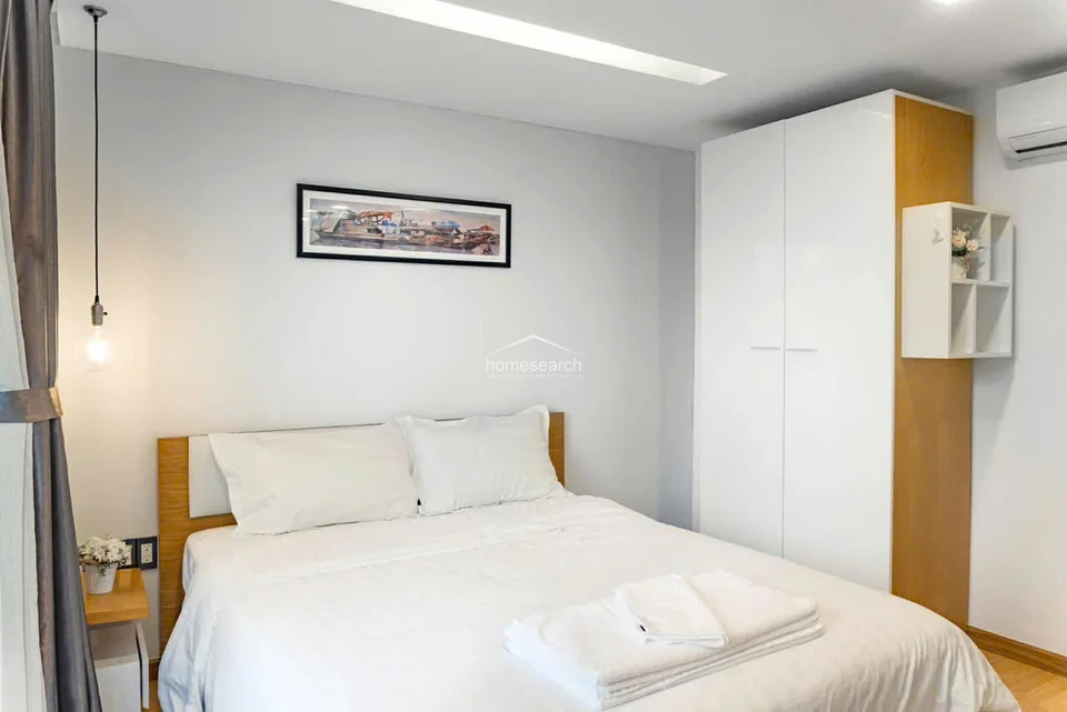 This bedroom provides a serene retreat with a comfortable double bed, white linens, and a tall, white wardrobe with integrated wood-finished shelving. The room is illuminated by a stylish pendant light and features light-toned wood flooring, creating a warm and well-maintained atmosphere.