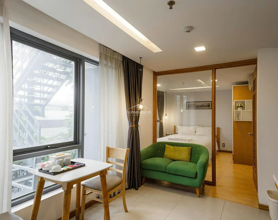 This living area features a modern, open-plan layout with a cozy green sofa and a small dining table positioned by a large window that offers natural light and a view of an outdoor metal staircase. The space is finished with neutral-toned floor tiles and a minimalist ceiling design, seamlessly connecting to the adjacent bedroom through a large glass partition framed in wood.