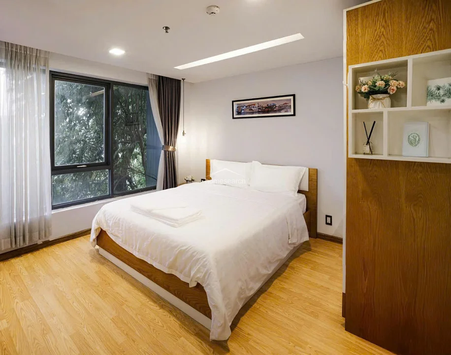 This bedroom layout highlights the connection between the sleeping area and the rest of the unit, emphasizing the bright, airy feel provided by the large window and sheer curtains. The room is well-organized, featuring a wooden bed frame, a decorative wall print, and an open shelving unit that adds both storage and a touch of modern decor.