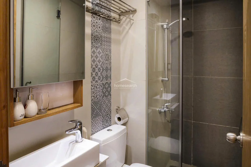The bathroom is compact yet modern, featuring a glass-enclosed shower with dark tile accents and a sleek white vanity with a mirror cabinet. The space is well-maintained, utilizing a mix of patterned and solid tiles to create a contemporary look while providing essential storage on the vanity shelf.