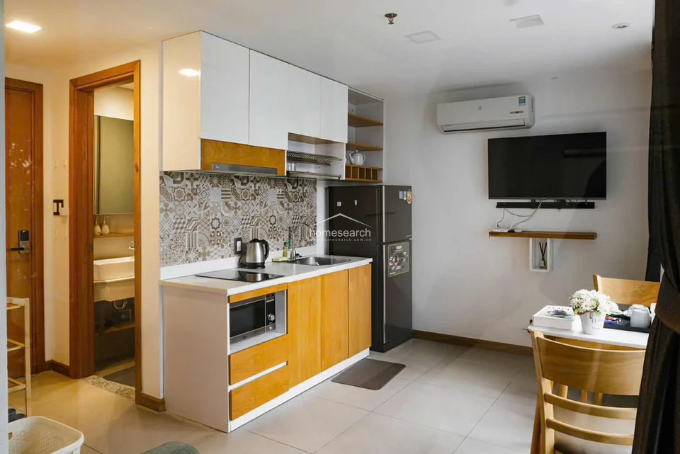The kitchenette is efficiently designed with white and wood-toned cabinetry, a patterned tile backsplash, and essential appliances including a built-in microwave, induction cooktop, and refrigerator. It maintains a clean, functional aesthetic with light-colored flooring and is equipped with a wall-mounted AC unit and television for added convenience.