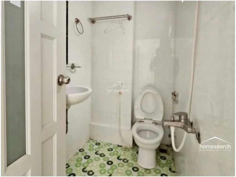 This compact bathroom is finished with white wall tiles and a distinctive green-patterned floor tile, creating a functional, utilitarian space. It is equipped with a wall-mounted sink, a standard toilet, and a handheld shower fixture, presenting a clean but basic level of maintenance.