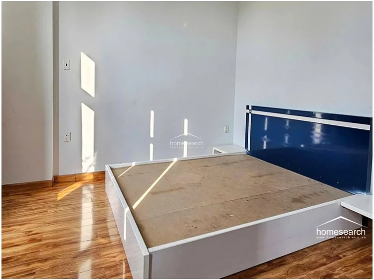 This bedroom is finished with light-colored walls, wood-look flooring, and a simple platform bed frame with a navy blue headboard. The room maintains a clean, minimalist aesthetic with basic electrical outlets and switches visible on the wall.