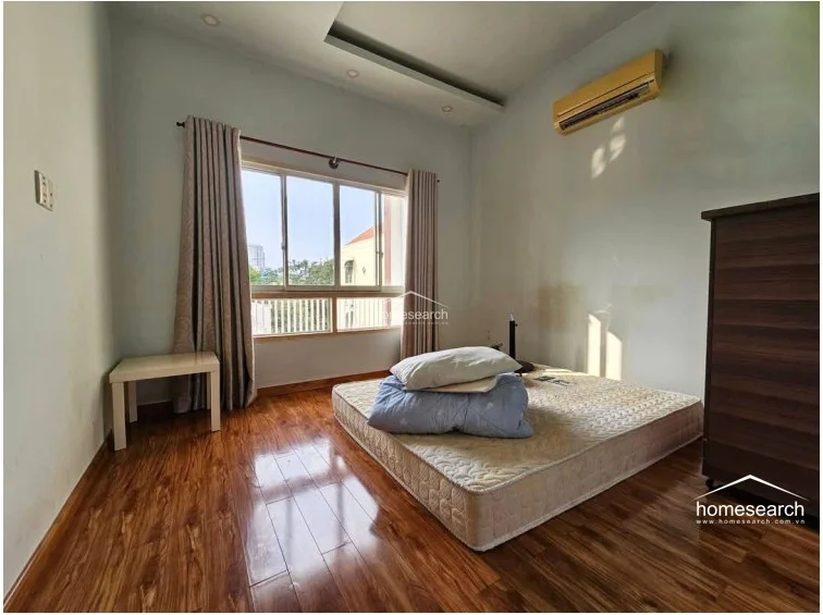 This bedroom is equipped with a wall-mounted air conditioning unit and features wood-look flooring throughout. A large window with neutral-toned curtains allows for ample natural light, while the room is furnished with a mattress on a low platform and a tall dark wood cabinet.