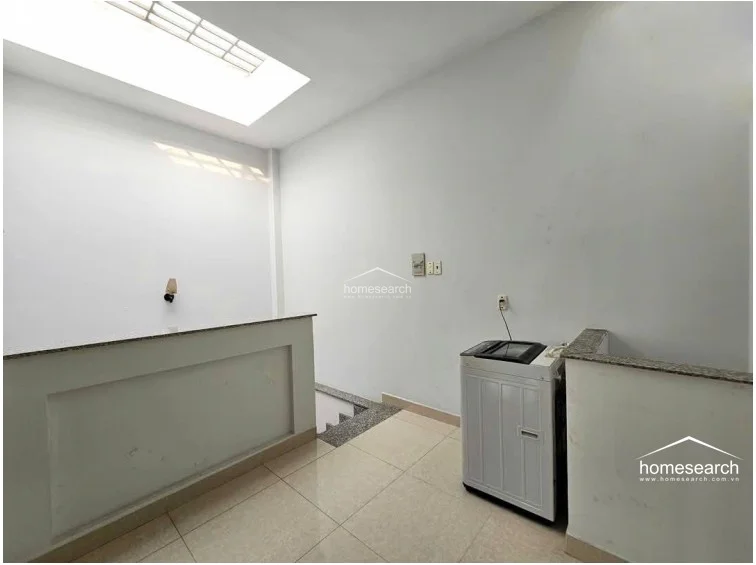 This utility area or landing features light-colored tile flooring and a top-loading washing machine placed near the stairwell. The space is illuminated by a large skylight above, creating a bright, open environment with white walls and a simple, functional layout.