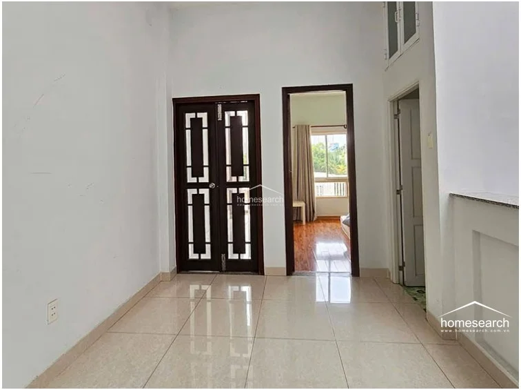 This landing or hallway area features neutral light-colored floor tiles and white walls, providing a bright and airy circulation space. It connects to a bedroom visible through the open doorway, which boasts contrasting warm-toned wood flooring and natural light from a window.
