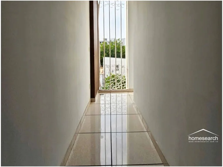 This narrow hallway or transition space is finished with light-colored floor tiles and plain white walls, creating a minimalist aesthetic. A tall, barred window at the end provides natural light and a view of the exterior, while the overall condition appears well-maintained.
