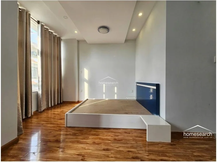 This bedroom features warm wood-look flooring and a minimalist platform bed frame with a navy blue headboard. The room is brightened by floor-to-ceiling curtains covering a large window, and it includes a modern recessed ceiling design with integrated lighting.