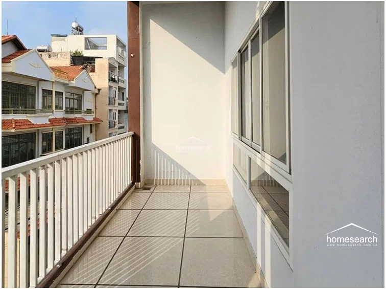 This outdoor balcony features light-colored tile flooring and a white metal safety railing, offering a view of the surrounding residential neighborhood. The space is accessible via large sliding glass windows, providing a clean and functional area for outdoor ventilation or relaxation.